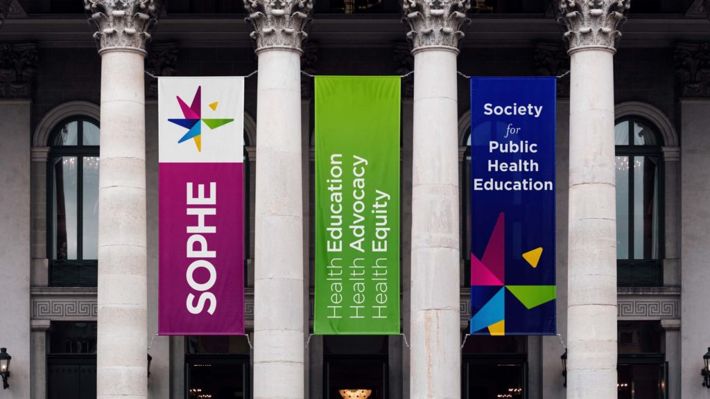 Here we see banners hanging between a building's pillars, showing SOPHE's new logo, tagline, and design.
